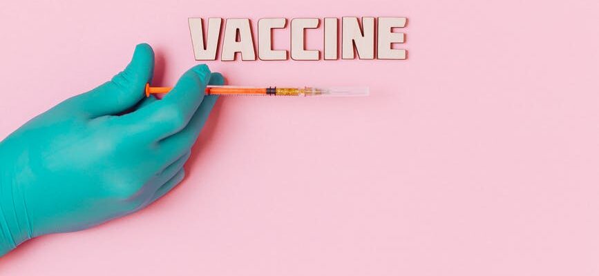vaccine text and a person wearing latex glove while holding a syringe on pink background